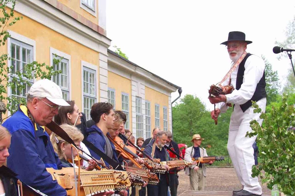 Hasse Gille gra na nyckelharpie na sceną pod którą stoją inni muzykujący na nyckelharpach uczestnicy festiwalu.