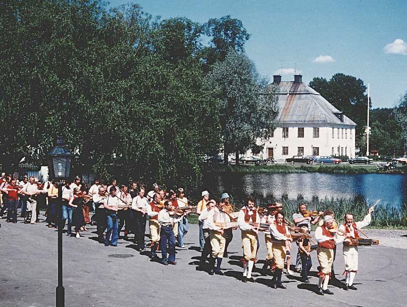 Pochód muzykantów podczas spelmastamma w Szwecji na tle rzeki i budynków w oddali
